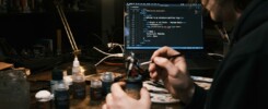 A mini painter sits painting a model with web code on a PC monitor in the background.