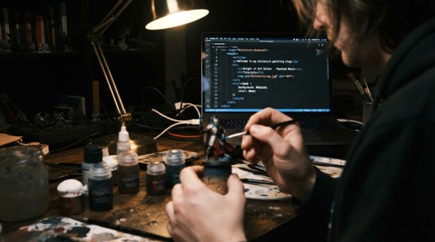 A mini painter sits painting a model with web code on a PC monitor in the background.